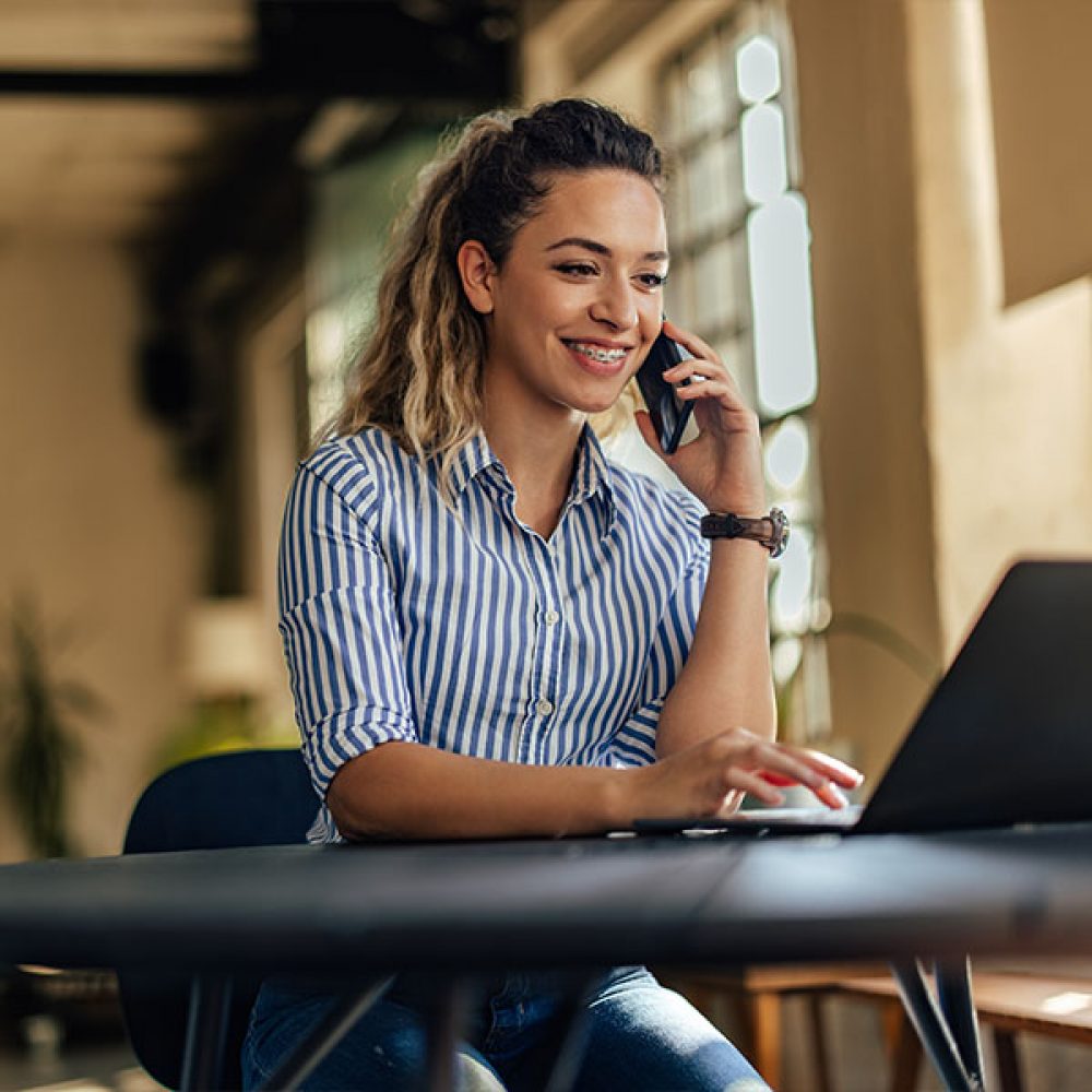 Smiling girl working over the laptop, making a phone call, talking to someone.