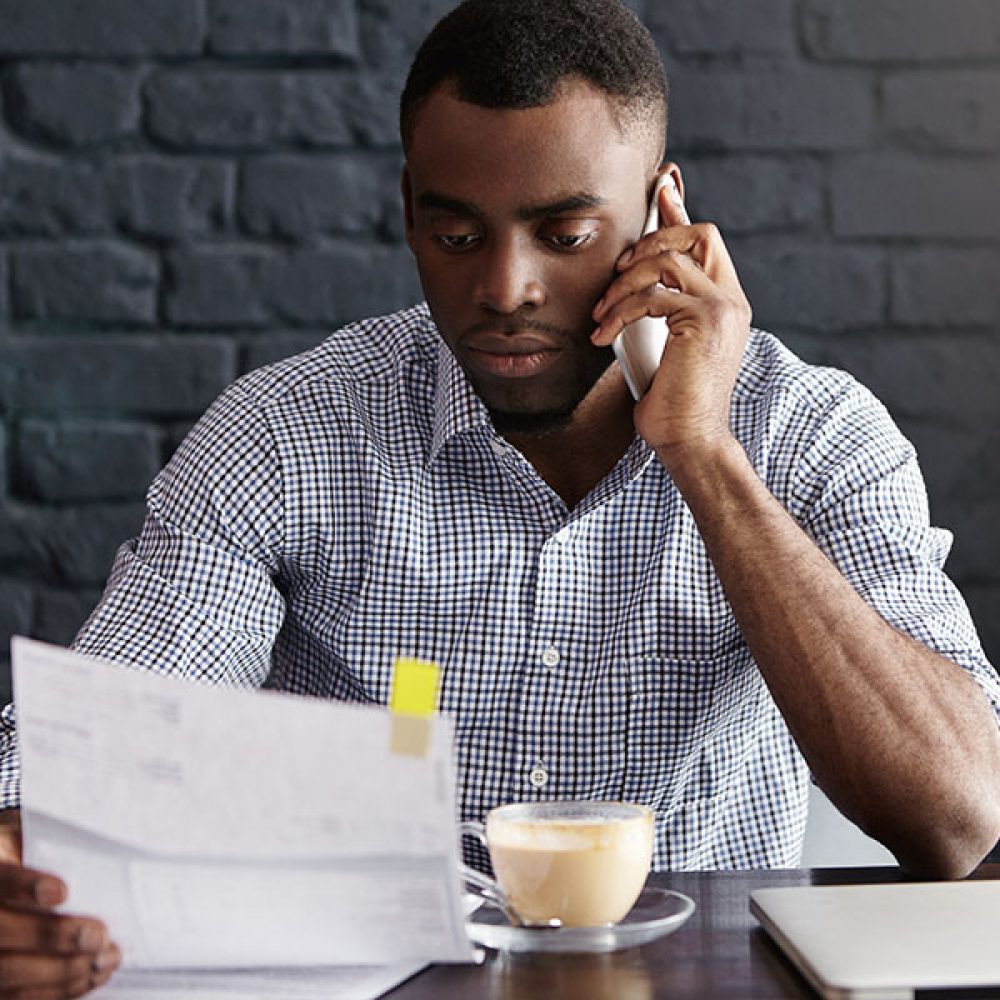 Serious African entrepreneur holding piece of paper in one hand and mobile phone in other, having conversation with someone, receiving unexpected negative news during coffee break at cafeteria